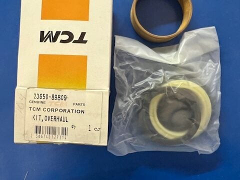 TCM genuine parts box labeled 'Kit, Overhaul' with a plastic bag containing a yellow seal and a circular cardboard spacer beside it on a blue surface