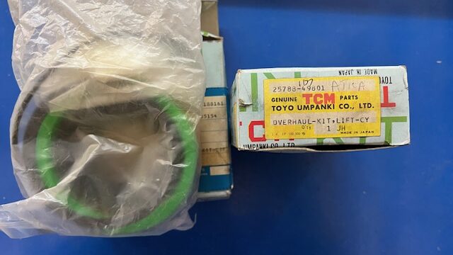 Green rubber seal in a clear plastic bag beside a small box labeled Genuine Toyota Parts Overhaul Kit on a blue surface.
