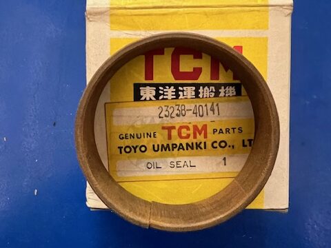 Brown oil seal placed over a yellow TCM genuine parts box, showcasing the packaging label beneath the seal (TOYO UMPANKI CO., LTD). Connects to automotive seals topic.