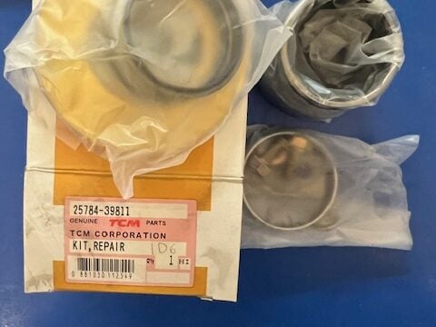Box labeled Genuine Parts TCM Corporation Kit Repair with two metal cylindrical parts in plastic bags on a blue surface.