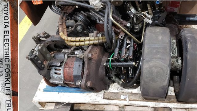 Forklift engine and drive belt assembly mounted on a wooden pallet, with a rusty motor and exposed wiring beside a labeled Toyota Electric Forklift plate on the left panel.
